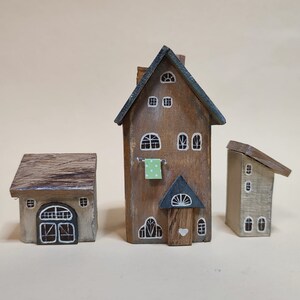 May include: Three miniature wooden houses with painted details. The largest house is brown with a green roof and white windows. The middle house is gray with a brown roof and a white door. The smallest house is beige with a brown roof and white windows.
