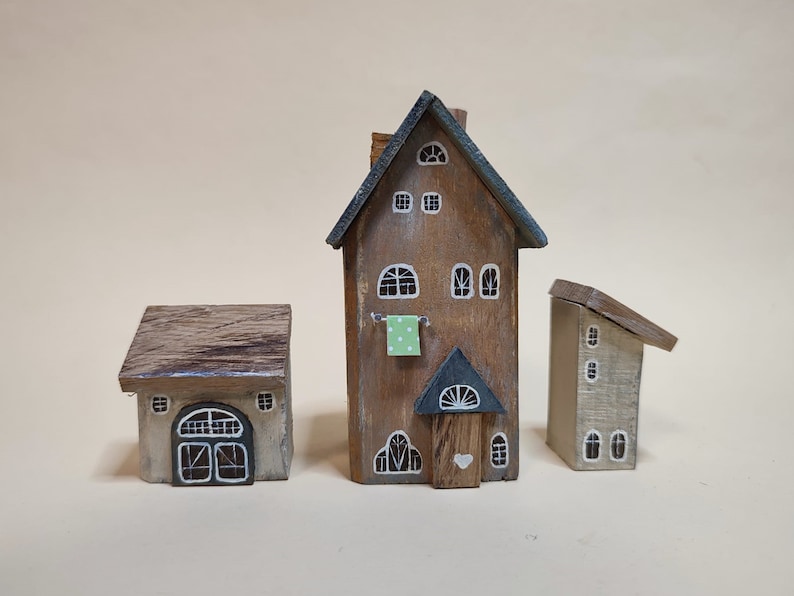 May include: Three miniature wooden houses with painted details. The largest house is brown with a green roof and white windows. The middle house is gray with a brown roof and a white door. The smallest house is beige with a brown roof and white windows.
