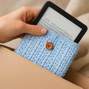 May include: A light blue knitted e-reader sleeve with a wooden button closure. The sleeve is holding a black e-reader with a white screen. The sleeve is inside a tan leather case.
