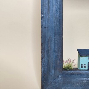 May include: A small, light blue house with black trim and windows sits in a wooden frame. The house is surrounded by a small patch of green grass.