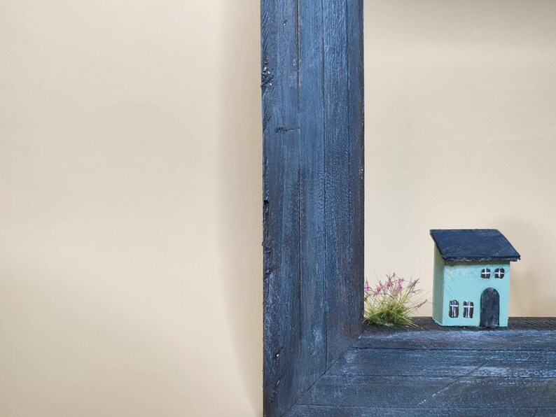 May include: A small, light blue house with black trim and windows sits in a wooden frame. The house is surrounded by a small patch of green grass.