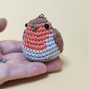 May include: A small, handmade crochet bird keychain. The bird features a round body with a red breast, gray and blue belly, and brown wings and head. It has small black eyes and a metal loop for attaching to keys or a bag.