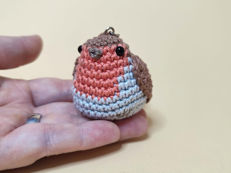 May include: A small, handmade crochet bird keychain. The bird features a round body with a red breast, gray and blue belly, and brown wings and head. It has small black eyes and a metal loop for attaching to keys or a bag.