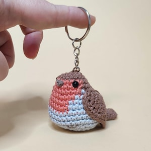 May include: A handmade crochet keychain featuring a small, round bird. The bird has a red breast, brown wings and back, and a light blue belly. It is attached to a silver-colored keyring.