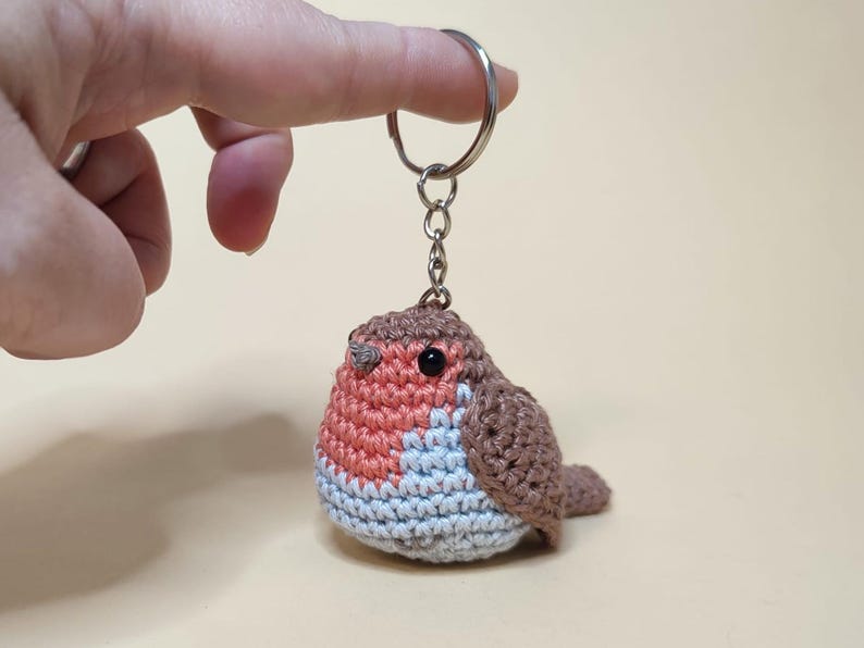 May include: A handmade crochet keychain featuring a small, round bird. The bird has a red breast, brown wings and back, and a light blue belly. It is attached to a silver-colored keyring.