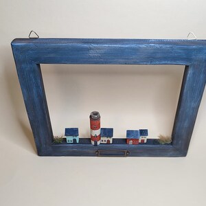 May include: A wooden frame with a blue painted finish, featuring a miniature lighthouse and houses. The frame has a metal hanger and a small metal nameplate.