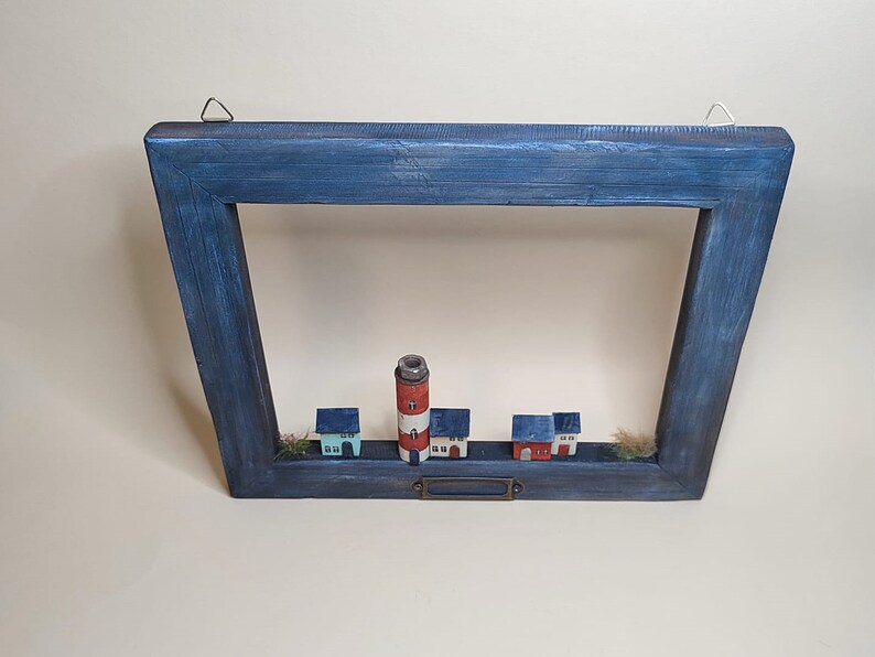 May include: A wooden frame with a blue painted finish, featuring a miniature lighthouse and houses. The frame has a metal hanger and a small metal nameplate.
