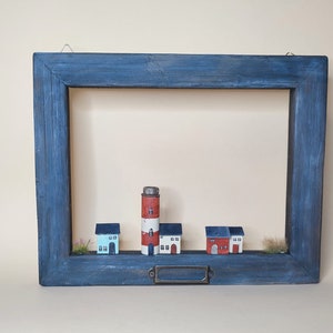 May include: A blue wooden frame with a white and red lighthouse and four small houses inside. The houses are white with red roofs and windows. The frame has a metal hook on the top and a metal nameplate on the bottom.