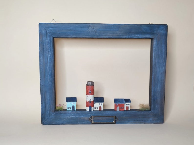 May include: A blue wooden frame with a white and red lighthouse and four small houses inside. The houses are white with red roofs and windows. The frame has a metal hook on the top and a metal nameplate on the bottom.