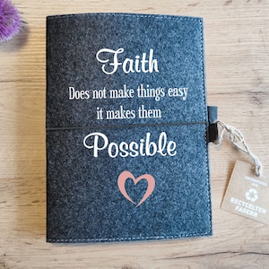 May include: A gray felt notebook with the text "Faith does not make things easy it makes them Possible" and a pink heart. The notebook has a brown tag with the text "Recycelten Fasern" and a recycling symbol.