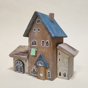 May include: A wooden miniature house with a blue roof, a green door, and white painted windows. The house has a small, attached building with a brown roof and a white door. The house is decorated with a green and white patterned cloth hanging on a clothesline.