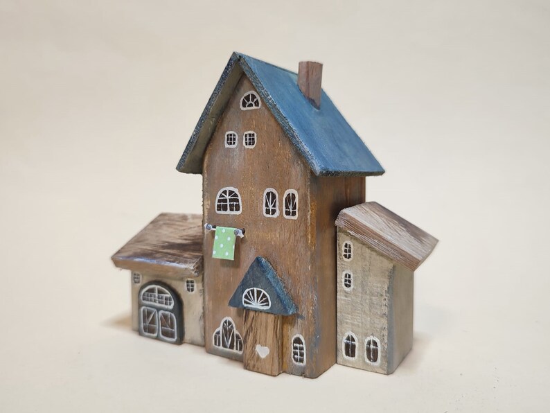 May include: A wooden miniature house with a blue roof, a green door, and white painted windows. The house has a small, attached building with a brown roof and a white door. The house is decorated with a green and white patterned cloth hanging on a clothesline.