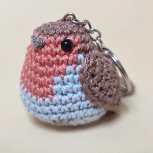 May include: A handmade crochet keychain in the shape of a robin bird. The bird features a red breast, light blue belly, and brown head and wings. It has a black eye and a silver keychain ring.