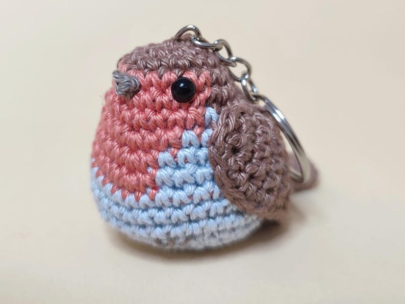 May include: A handmade crochet keychain in the shape of a robin bird. The bird features a red breast, light blue belly, and brown head and wings. It has a black eye and a silver keychain ring.