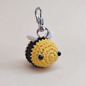 May include: A crocheted yellow and black bee keychain with white wings and black bead eyes. The bee is attached to a silver metal key ring.