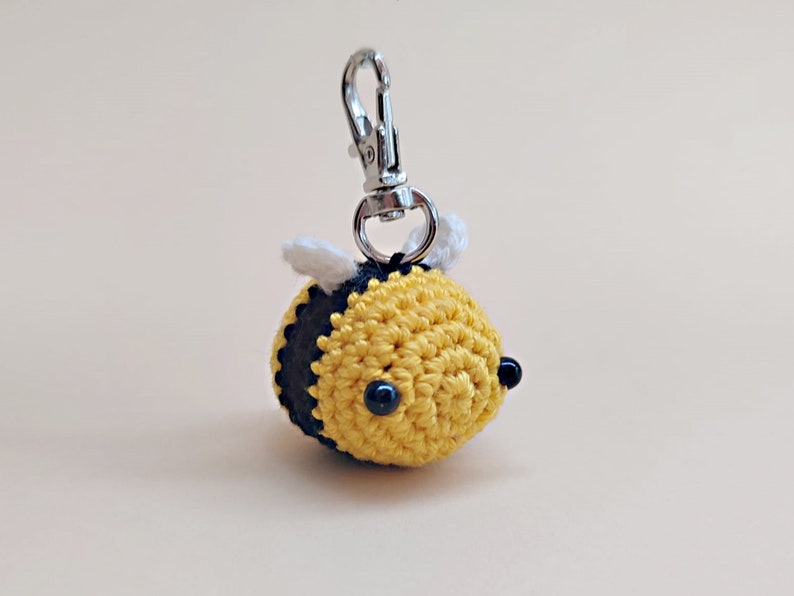 May include: A crocheted yellow and black bee keychain with white wings and black bead eyes. The bee is attached to a silver metal key ring.