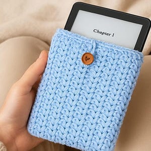 May include: A light blue, hand-knitted e-reader case with a wooden button closure. The case holds a black e-reader displaying the text "Chapter 1". The case is made of a soft, textured yarn.