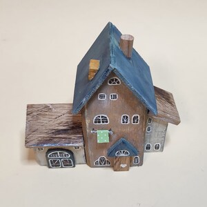 May include: A miniature wooden house with a blue roof, brown siding, and white windows. The house has a small porch with a green door and a white heart.