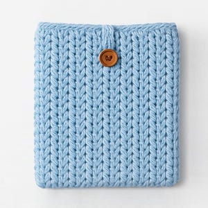May include: A light blue knitted tablet sleeve with a button closure. The sleeve is made of a thick, textured yarn and features a large, round, brown button. The knitted pattern is vertical and creates a ribbed effect.