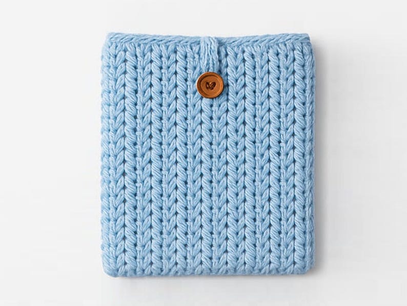 May include: A light blue knitted tablet sleeve with a button closure. The sleeve is made of a thick, textured yarn and features a large, round, brown button. The knitted pattern is vertical and creates a ribbed effect.