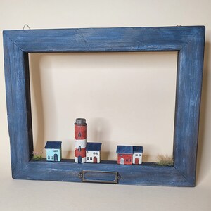 May include: A blue wooden frame with a white painted interior, featuring a miniature lighthouse and houses. The frame has a metal nameplate.