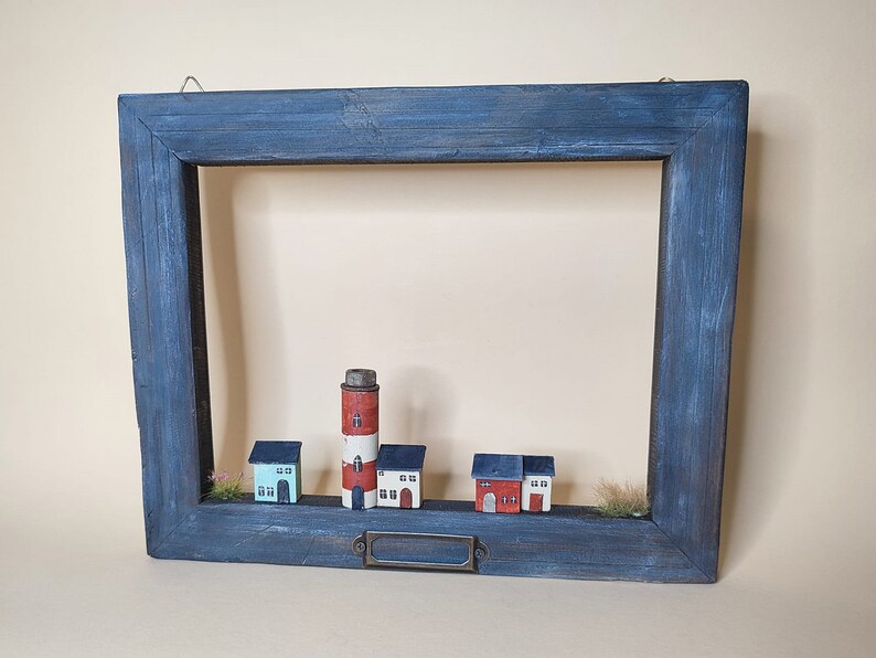 May include: A blue wooden frame with a white painted interior, featuring a miniature lighthouse and houses. The frame has a metal nameplate.