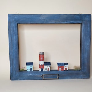 May include: A blue wooden frame with a miniature village scene inside. The scene includes a red and white lighthouse, three white houses with red roofs, and green grass.