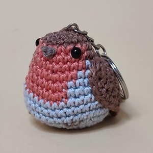May include: A handmade crochet robin keychain. The bird features a red breast, blue belly, and brown wings and head. It has black eyes and a silver keychain ring. The robin is a charming, whimsical accessory.