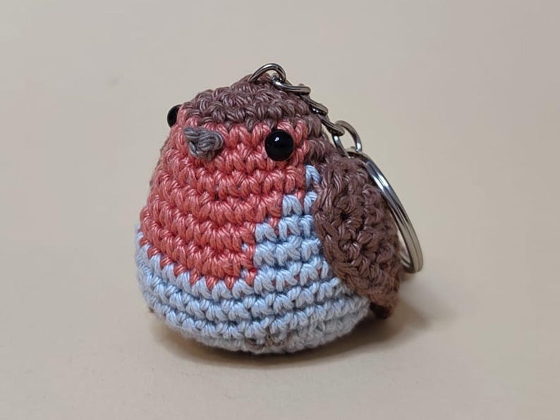 May include: A handmade crochet robin keychain. The bird features a red breast, blue belly, and brown wings and head. It has black eyes and a silver keychain ring. The robin is a charming, whimsical accessory.
