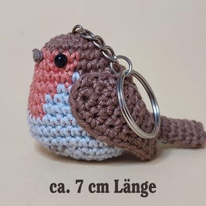 May include: Crocheted bird keychain, approximately 4 cm high and 7 cm long. The bird features a brown body, a pink chest, and a light blue belly. It has a silver keychain ring attached.