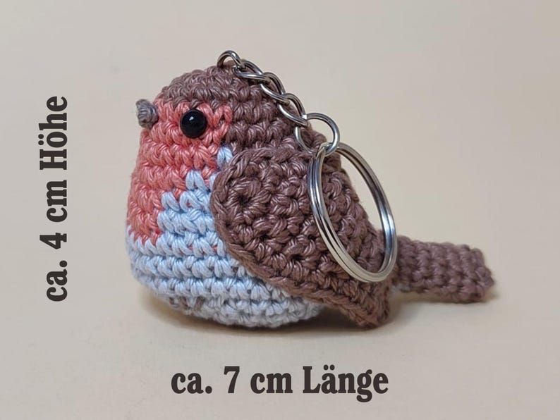 May include: Crocheted bird keychain, approximately 4 cm high and 7 cm long. The bird features a brown body, a pink chest, and a light blue belly. It has a silver keychain ring attached.