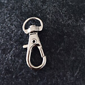 May include: Silver metal swivel clasp hook with a spring-loaded gate. The hook is designed for attaching to keychains, bags, or other items.