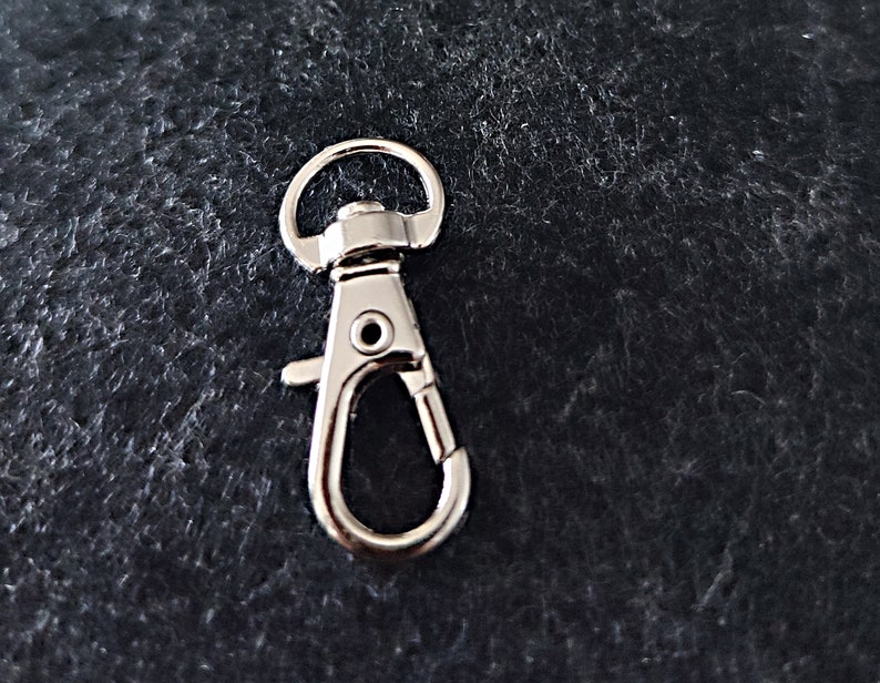 May include: Silver metal swivel clasp hook with a spring-loaded gate. The hook is designed for attaching to keychains, bags, or other items.