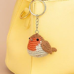 May include: A handmade crochet bird keychain with a silver keyring and chain. The bird has a brown, orange, and white color scheme. The keychain is hanging in front of a yellow handbag.