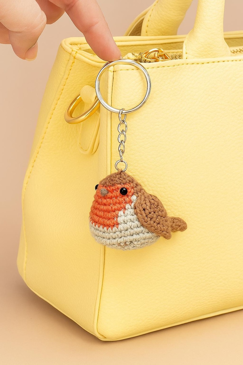 May include: A handmade crochet bird keychain with a silver keyring and chain. The bird has a brown, orange, and white color scheme. The keychain is hanging in front of a yellow handbag.