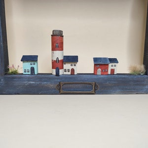 May include: A wooden frame with a blue painted finish displays a miniature village scene. The scene includes a red and white lighthouse, a blue and white house, a white house with a red roof, and a red house with a white roof. The houses are all made of wood and have small windows and doors.