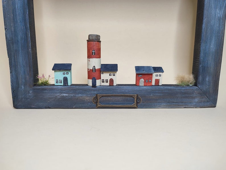 May include: A wooden frame with a blue painted finish displays a miniature village scene. The scene includes a red and white lighthouse, a blue and white house, a white house with a red roof, and a red house with a white roof. The houses are all made of wood and have small windows and doors.