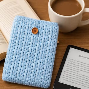 May include: A light blue knitted e-reader cover with a wooden button closure. The cover is next to an open book, a cup of coffee, and an e-reader displaying the text "Churper 20".