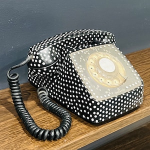 May include: A black rotary phone with white polka dots. The phone has a black rotary dial with white numbers and letters. The phone has a black coiled cord.