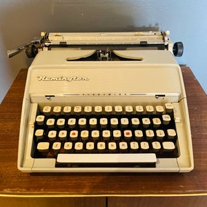 May include: A vintage white Remington  Fleetwing typewriter with a black keyboard and red keys. The typewriter has a black carriage return lever and a black tab key.