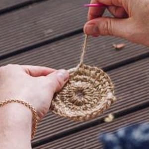 May include: A person is crocheting a round, brown, jute coaster. The coaster has a raised centre and is being worked on with a pink crochet hook.