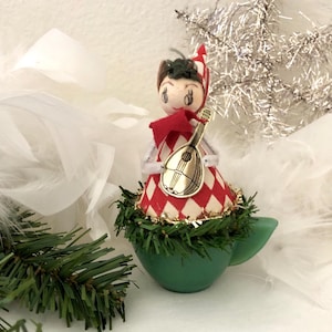 May include: A vintage Christmas ornament featuring a doll-like figure in a red and white diamond pattern dress, holding a golden lute. The figure sits atop a green teacup-shaped base, surrounded by greenery and white feathers. A silver star adds a festive touch.