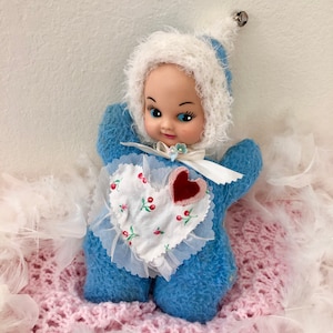 May include: A blue plush baby doll with a white crocheted hat and a heart-shaped patch on its chest. The doll is sitting on a pink and white blanket.