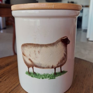 May include: White ceramic canister with a wooden lid and a brown and white sheep illustration. The sheep is standing on a green patch of grass.