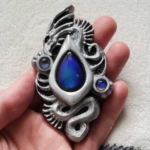 May include: A silver pendant featuring a dragon design with a blue iridescent gemstone in the center and two smaller blue gemstones on the sides.