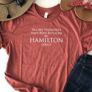 May include: A red t-shirt with white text that reads "All my thoughts have been replaced by Hamilton Lyrics".