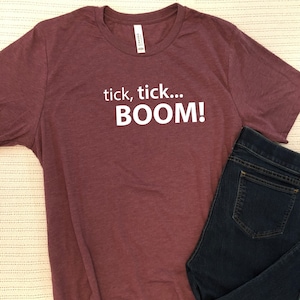 May include: A maroon t-shirt with the text "tick, tick... BOOM!" printed in white.