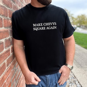 May include: Black t-shirt with the white text "MAKE CHEVYS SQUARE AGAIN." The shirt is being worn by a person leaning against a brick wall. The person is wearing blue jeans and a watch.