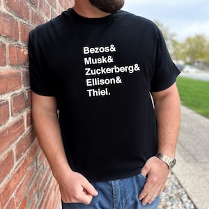 May include: Black t-shirt with white text listing names: Bezos, Musk, Zuckerberg, Ellison, and Thiel. The person wearing the shirt is leaning against a red brick wall, wearing blue jeans and a watch.