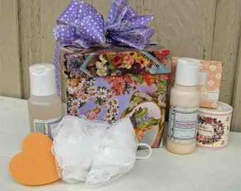 May include: A gift set with a floral-decorated box tied with a purple polka dot bow. The set includes bottles, a heart-shaped sponge, a loofah, and a candle. The bottles have white caps and labels. The candle is in a floral-decorated container.